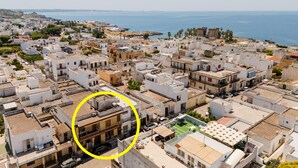 Une ville côtière aux bâtiments blancs, offrant une vue dégagée sur la mer, et dont l’un des bâtiments se distingue par la présence d’un balcon.