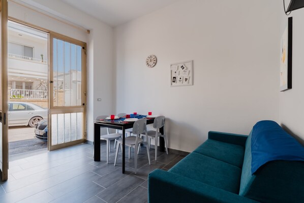 A modern living room with a teal sofa, a dining table with chairs, a sliding glass door leading outside, and a wall clock.