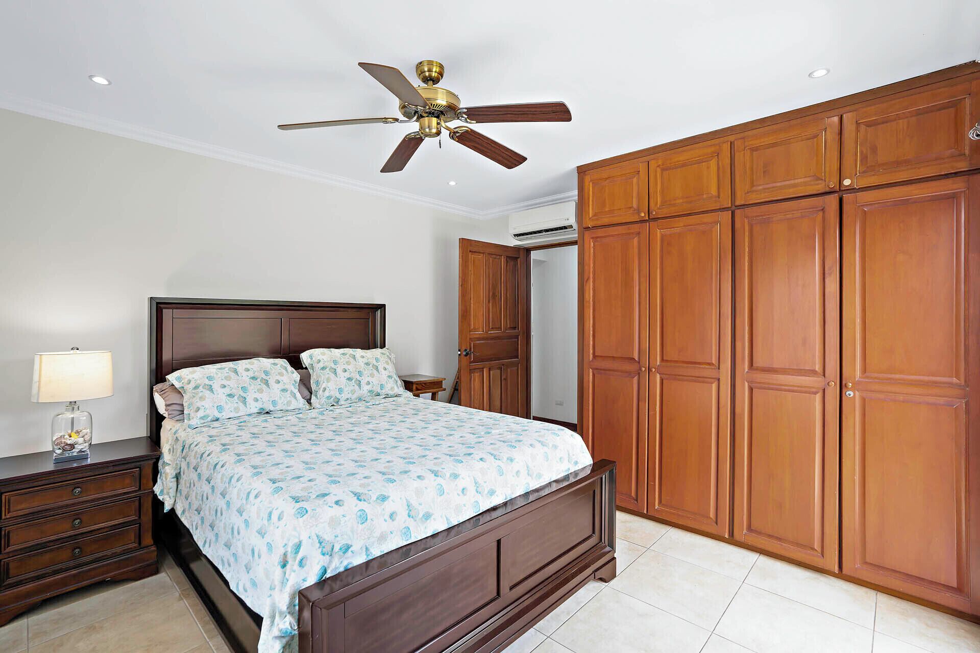 A bedroom with a bed, nightstand, lamp, wooden wardrobe, and ceiling fan.