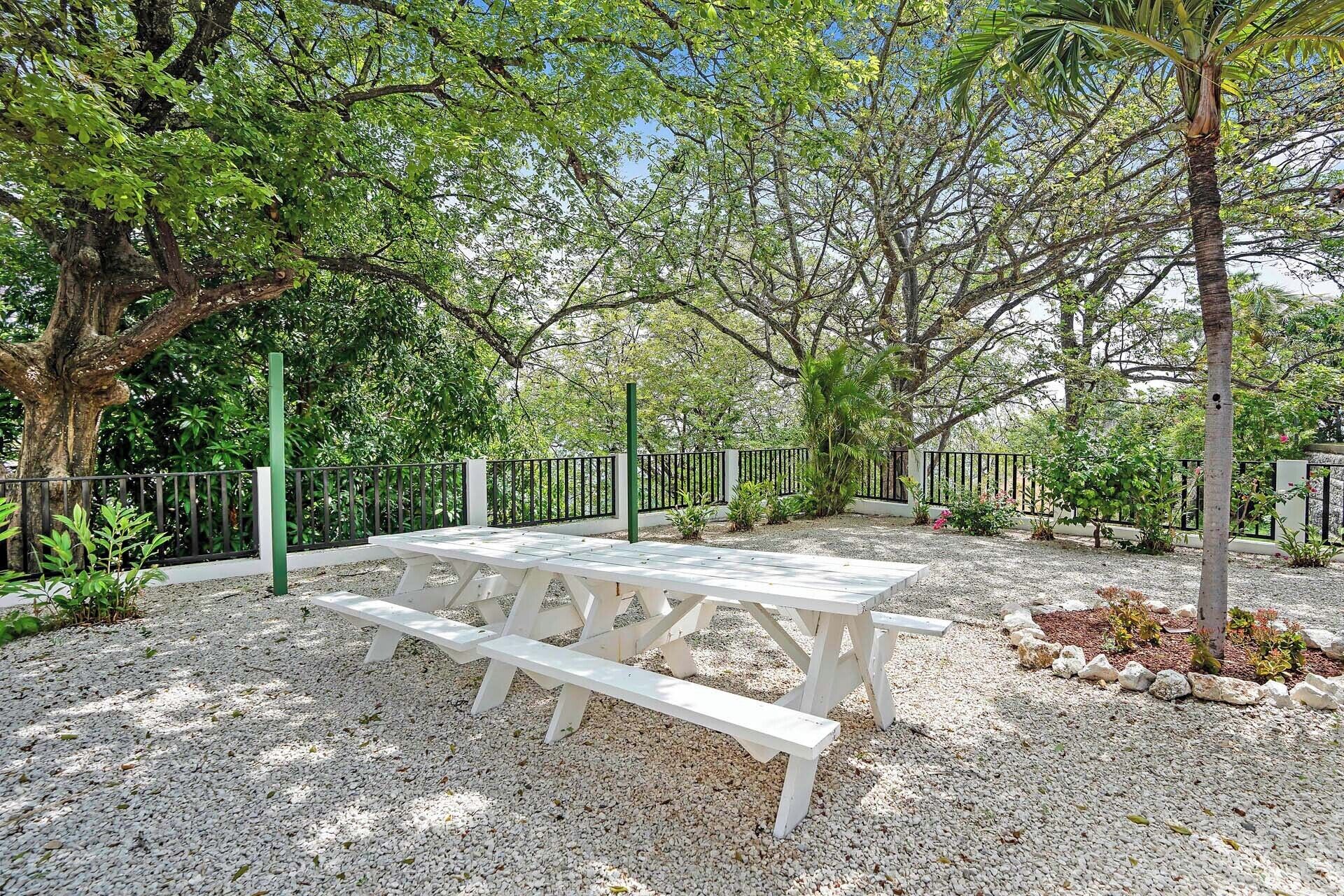Un área con mesas de picnic y bancos al aire libre, rodeada por un camino de grava y una valla metálica.