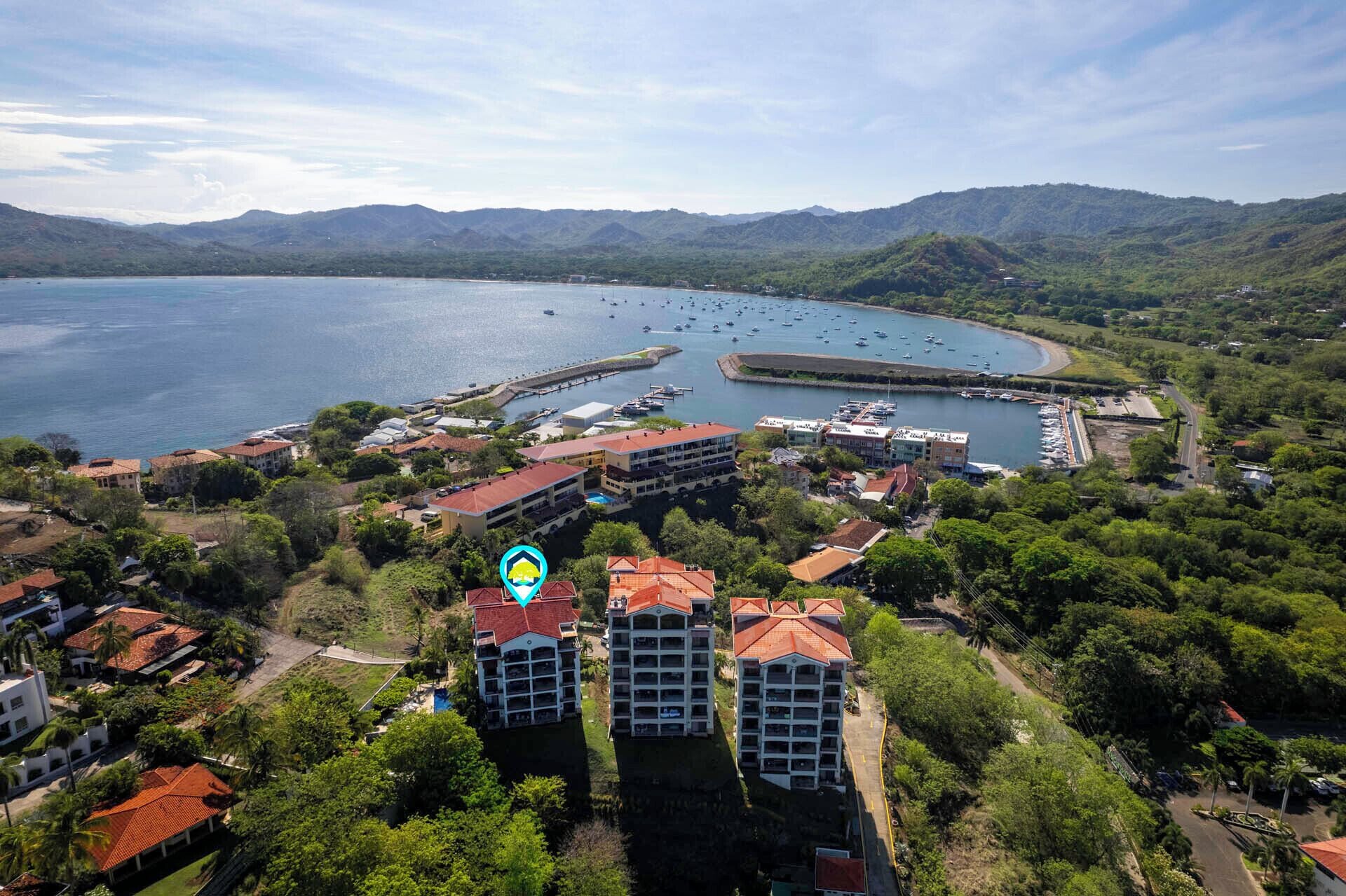 An aerial view of a coastal town with a marina, buildings with red roofs, and a mountainous backdrop.