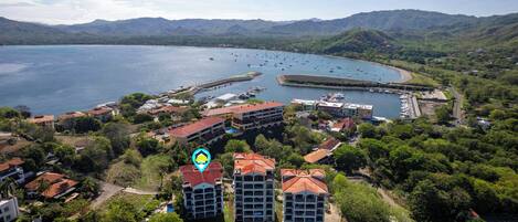 An aerial view of a coastal town with a marina, buildings with red roofs, and a mountainous backdrop.