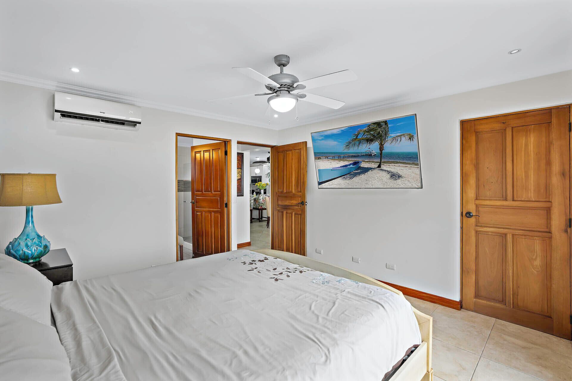 Une chambre à coucher avec un lit, un ventilateur de plafond, un appareil de climatisation, des portes en bois et un tableau sur le thème de la plage.