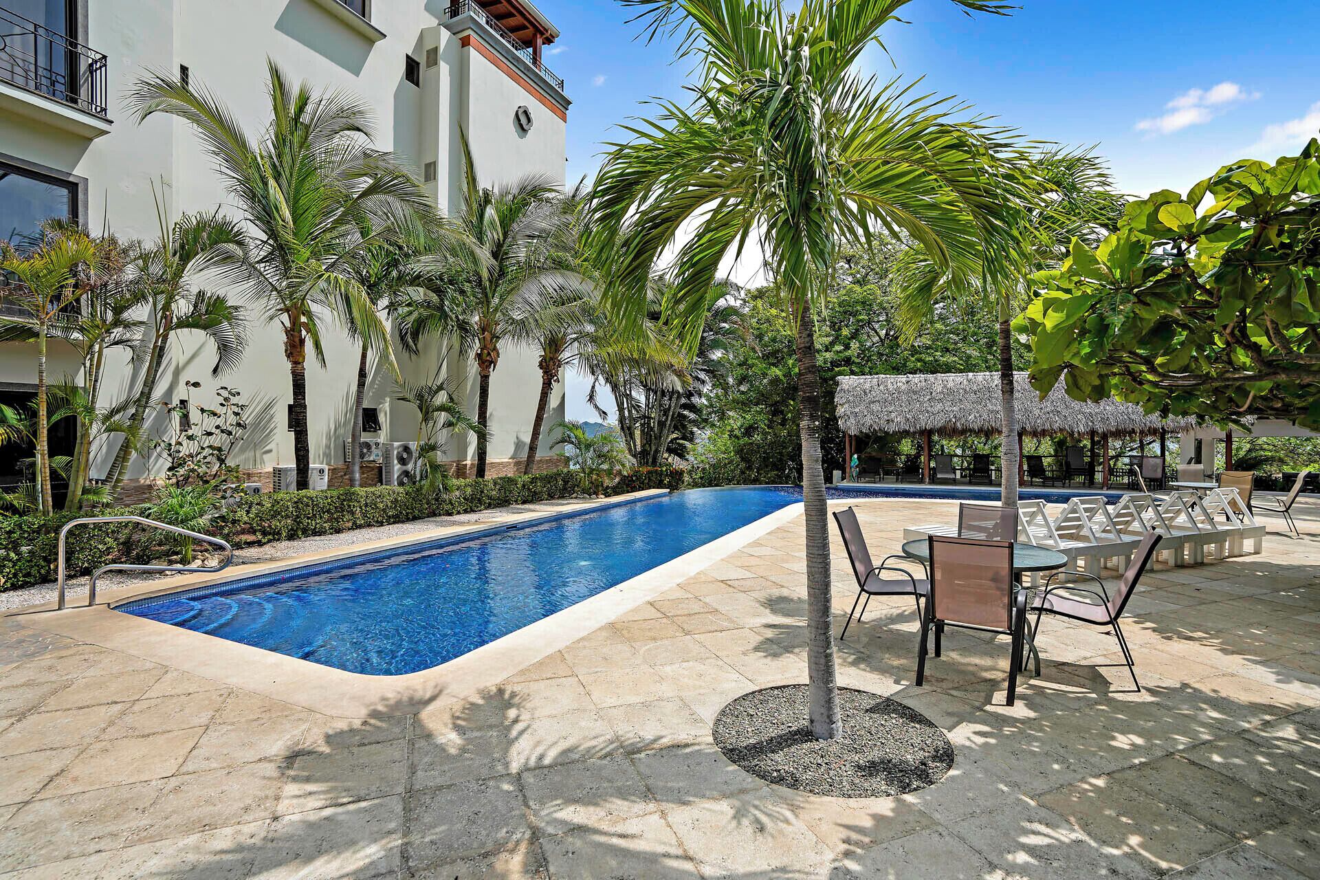 A swimming pool with lounge chairs and a thatched-roof structure, surrounded by palm trees and a building with a balcony.