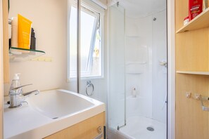 Un baño con lavamanos blanco, cabina de ducha de vidrio y estante con artículos de aseo.