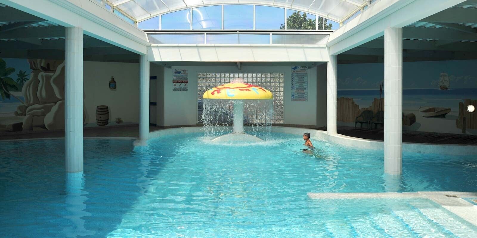 Une piscine intérieure dotée d’un élément décoratif aquatique au centre, entourée de murs ornés de peintures murales et de colonnes.