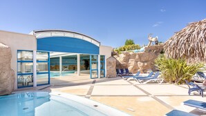 Un área de piscina con sillones de descanso, un edificio de fachada azul y una estructura con techo de paja.