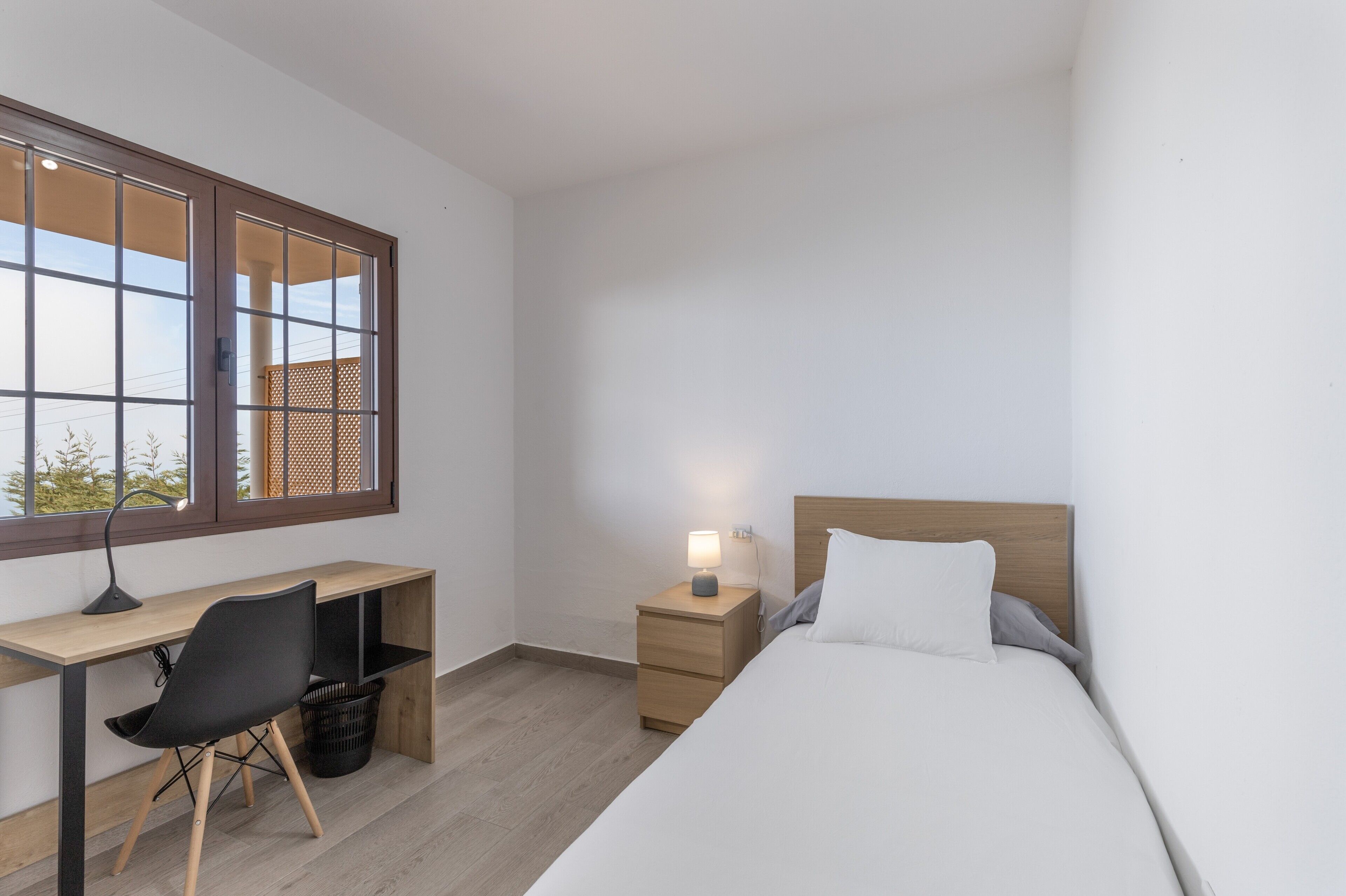 A bedroom with a bed, a desk, a chair, a nightstand with a lamp, and a window.
