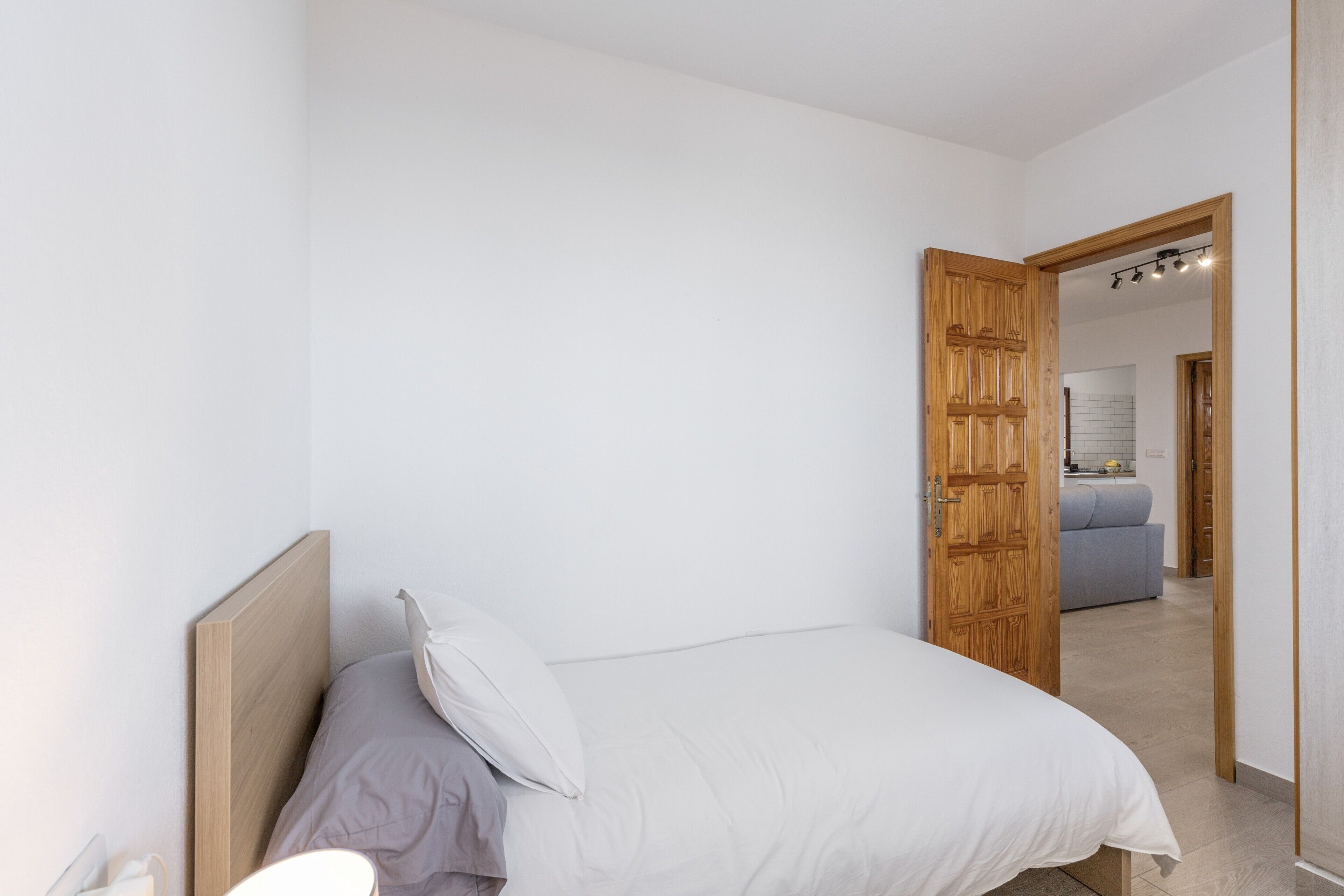 Une chambre à coucher avec une tête de lit en bois, un lit recouvert de linge de lit blanc et une porte ouverte donnant sur une salle de bain équipée d’un miroir et d’un lavabo.