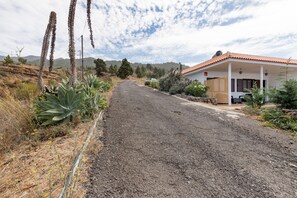 Un camino rural que lleva a un edificio blanco con techo de tejas rojas, rodeado de vegetación seca y montañas al fondo.