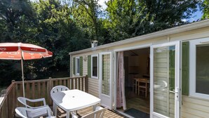 A wooden deck with a table and chairs, a covered umbrella, and a sliding glass door leading to an interior space. - Mobile Home 'Le Chataignier' with Shared Pool, Private Terrace and Private Garden (Saint-Pompon)
