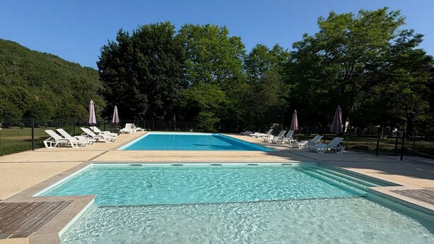 A rectangular swimming pool with clear water, surrounded by lounge chairs and umbrellas, set against a backdrop of trees and hills. - Mobile Home 'Le Chataignier' with Shared Pool, Private Terrace and Private Garden (Saint-Pompon)