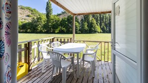 Ein überdachter Außenbereich mit Tisch und Stühlen, einer Holzterrasse und Blick auf ein Wiesenfeld mit Bäumen.