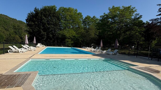 Ein Swimmingpool mit klarem Wasser, umgeben von Liegestühlen und Sonnenschirmen, vor einer Kulisse aus Bäumen und blauem Himmel.