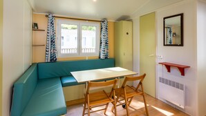 A compact room with a bench, a table, two chairs, a radiator, a window with curtains, and a mirror on the wall. - Mobile Home 'Le Chene' with Shared Pool, Private Terrace and Private Garden (Saint-Pompon)
