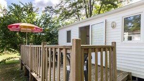 A mobile home with a wooden deck, a red and yellow umbrella, and a window with a view of trees. - Mobile Home 'Le Chene' with Shared Pool, Private Terrace and Private Garden (Saint-Pompon)