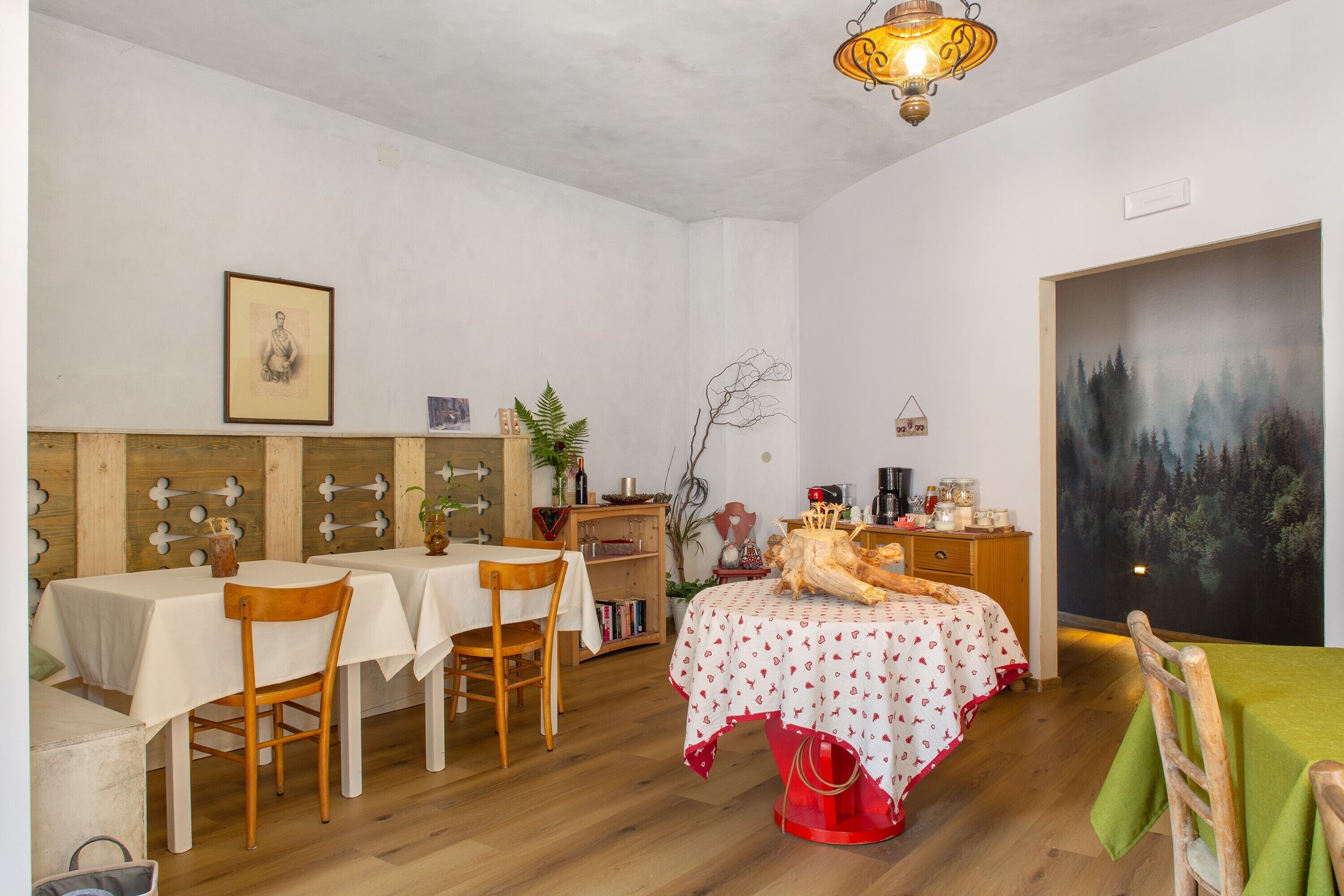 A dining area with tables and chairs, a bed with a red trunk, a painting of a forest, and a wooden wall with cut-out shapes.