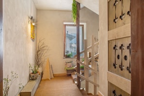 Un couloir étroit avec un escalier en bois, une fenêtre donnant sur un immeuble et une étagère fixée au mur, ornée de plantes.