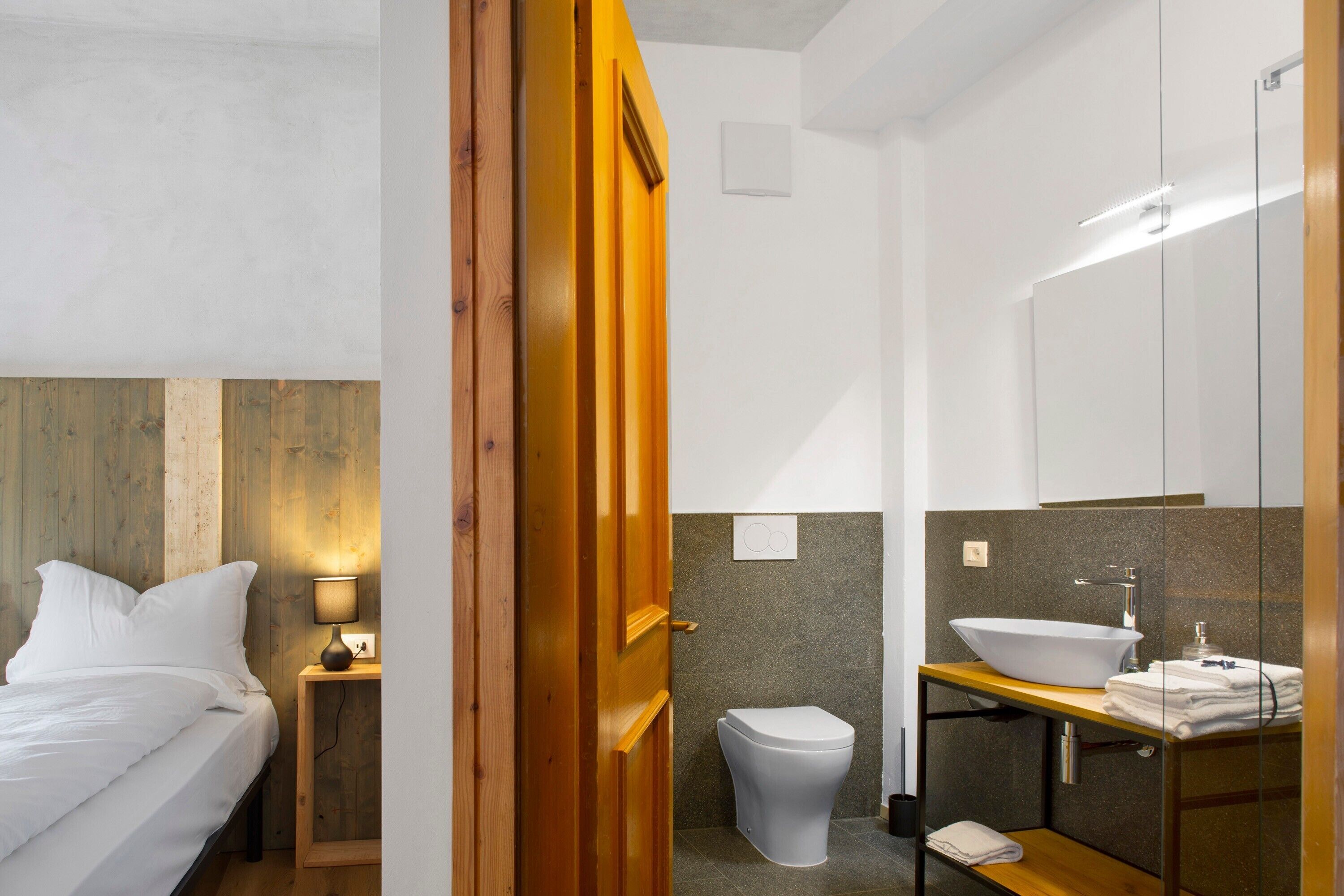 A hotel room with a bed, bedside table, lamp, wooden headboard, and a bathroom with a toilet, sink, mirror, and towel rack.