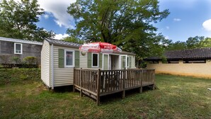 A small, single-story mobile home with a wooden deck and a red and white awning. - Mobile Home 'Le Noyer' with Shared Pool, Private Terrace and Private Garden (Saint-Pompon)