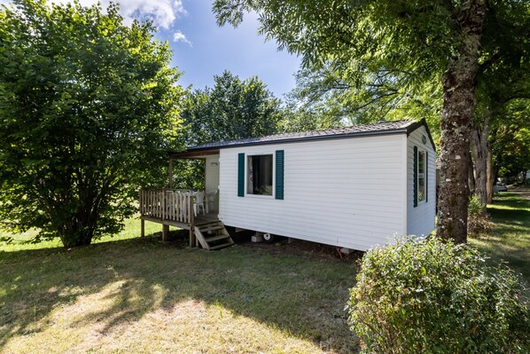 A small white mobile home with green shutters, a wooden deck, and a tree in the background. - Mobile Home 'Du Soleil' with Shared Pool, Private Terrace and Wi-Fi (Saint-Pompon)