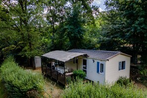 A small, single-story cabin with a porch, surrounded by dense greenery. - Mobile Home 'Du Soleil' with Shared Pool, Private Terrace and Wi-Fi (Saint-Pompon)