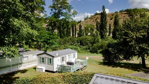 Une maison mobile dotée d’une véranda et d’une antenne parabolique, entourée de verdure et de collines.