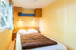 Ein kleines Zimmer mit einem Einzelbett, einem Holzkopfende, einer braunen Bettdecke und einem Fenster mit Vorhängen.