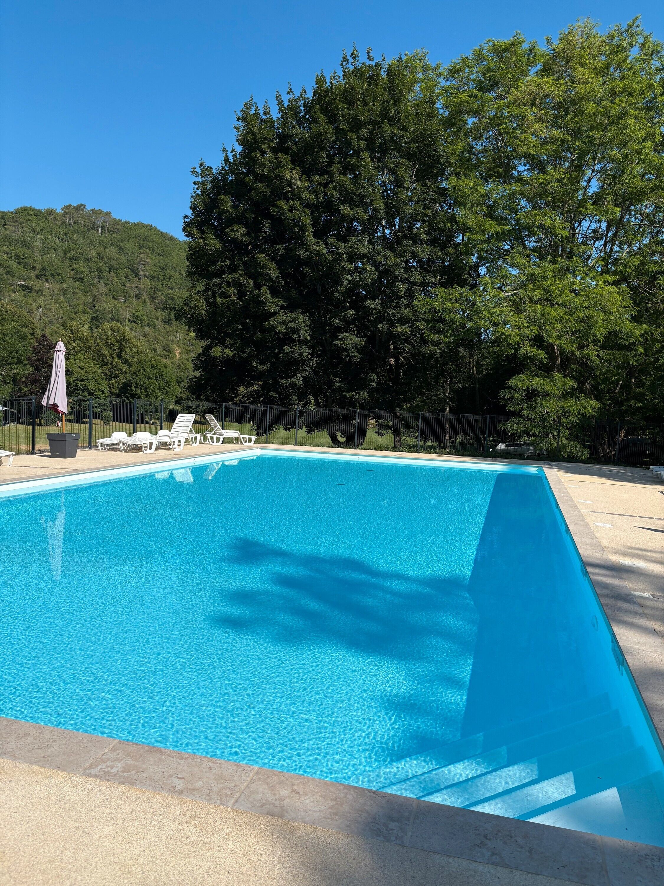 Une piscine rectangulaire à l’eau d’un bleu limpide, entourée d’une terrasse en béton et d’une clôture en métal.