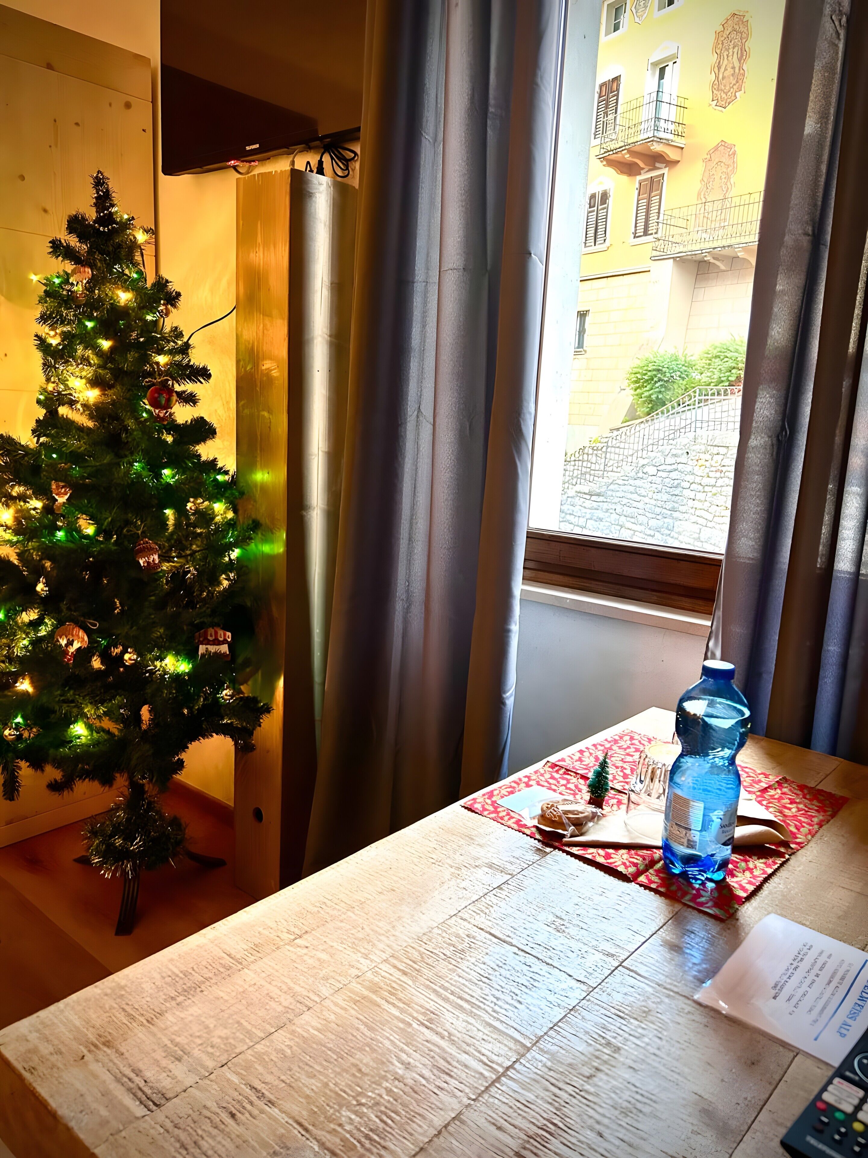 Un árbol de Navidad con luces, una botella de agua y un control remoto sobre una mesa de madera cerca de una ventana con cortinas.