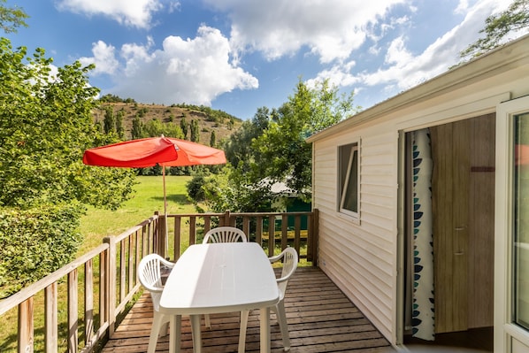 A wooden deck with a table and chairs, a red umbrella, and a view of a hillside.
