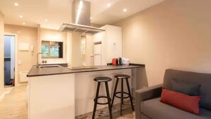 Una cocina moderna con una isla central, dos taburetes de bar y un sofá gris con un cojín rojo.