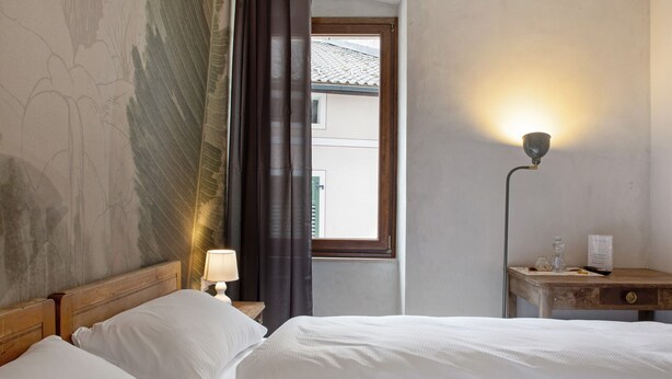 A hotel room with a bed, a nightstand with a lamp, a window with curtains, and a wall with a partial view of a building outside. - Room 'Con Sauna 2' with Mountain View and Wi-Fi (Castello Tesino)