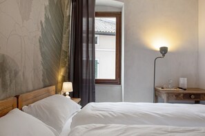 A hotel room with a bed, a nightstand with a lamp, a window with curtains, and a wall with a partial view of a building outside. - Room 'Con Sauna 2' with Mountain View and Wi-Fi (Castello Tesino)