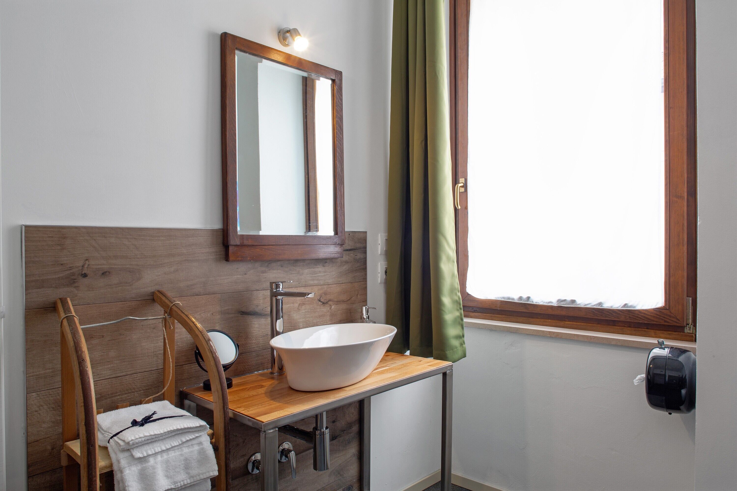 Un baño con tocador de madera, un lavamanos blanco, un espejo, una toalla, una ventana con cortinas y un dispensador de pañuelos.