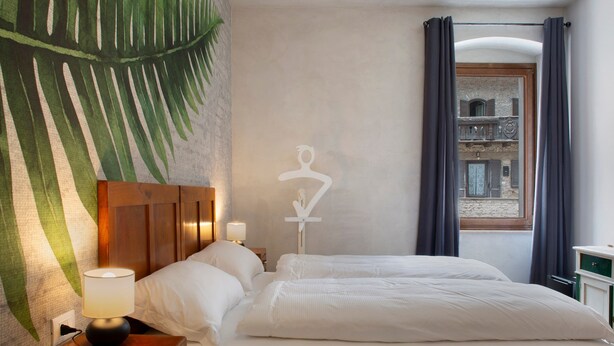 Ein Hotelzimmer mit einem Bett, einem Nachttisch mit Lampe, Wandgemälde, einem Fenster mit Vorhängen und Balkonblick.