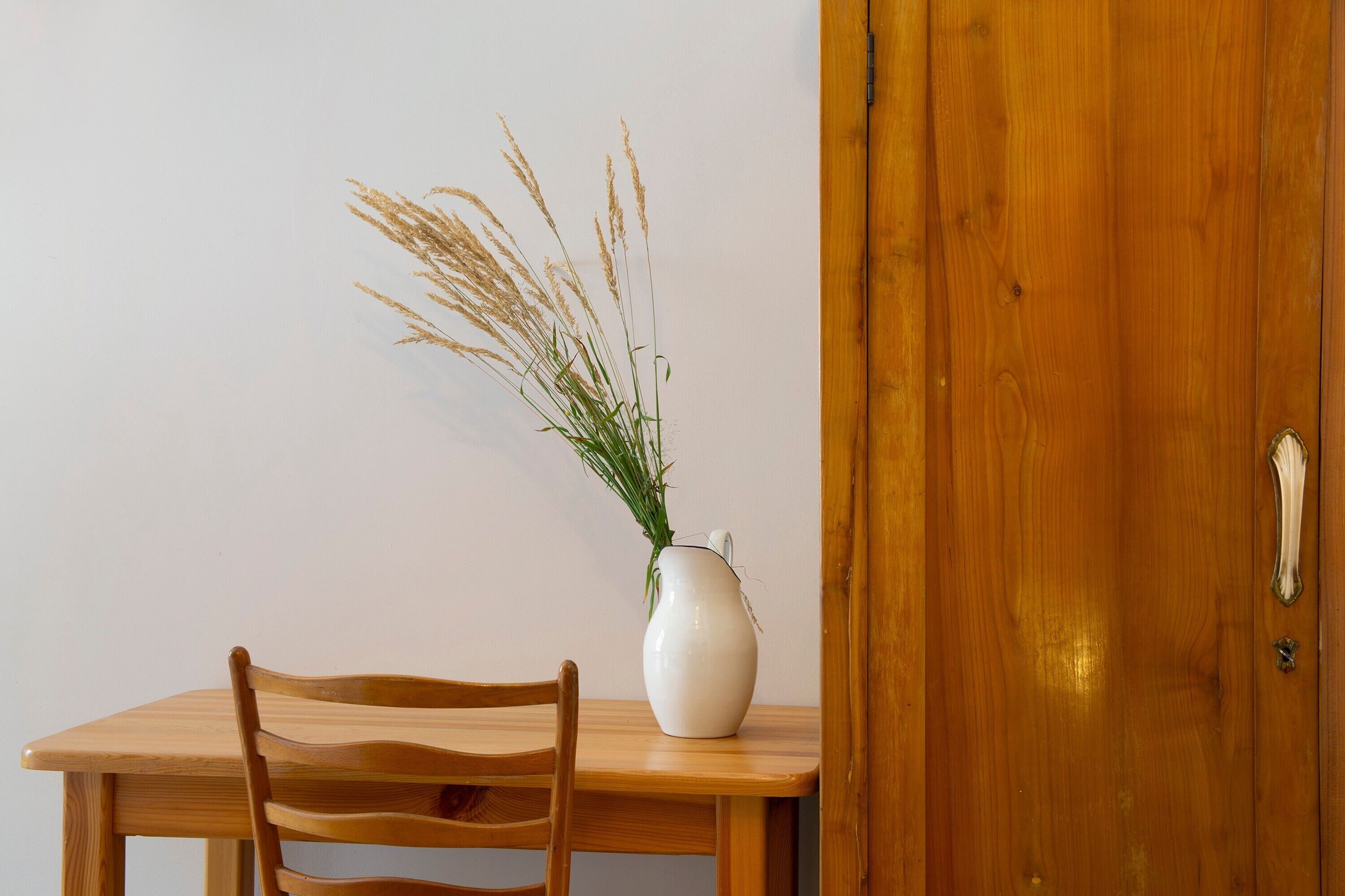 A wooden table with a white vase containing dried plants, a wooden chair, and a wooden door.