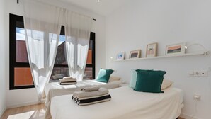 Ein Hotelzimmer mit zwei Betten, einem Fenster mit durchsichtigen Vorhängen, einem an der Wand befestigten Regal mit Bildern und einer sichtbaren Steckdose.