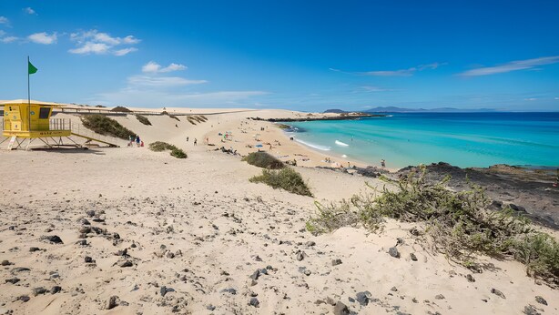 Ein Strand mit einem Rettungsturm, klarem türkisblauem Wasser und Sanddünen.