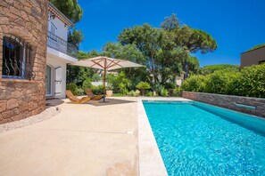 A swimming pool with a stone wall and a wooden deck with lounge chairs and an umbrella.