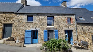 A stone building with two windows and a door, a small garden area with plants, and a bench.
