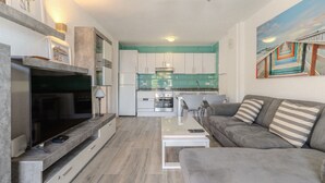 A modern kitchen with white cabinets, a dining area with a gray sofa, and a TV on a stand. - Vacation 'Colina Blanca Sunset View' with Sea View, Shared Pool and Wi-Fi (Costa Adeje)