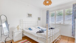 Een wit hemelbed met hoofdeinde, een nachtkastje met lamp, een hanglamp en een raam met gordijnen.