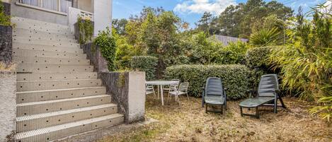 Una casa amb una escala enrajolada que condueix a un porxo cobert, una taula amb caires, i un jardí amb diverses plantes i arbres.