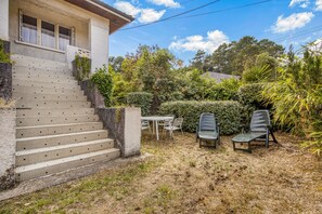 Une maison avec un escalier carrelé menant à un porche abrité, une table avec des chaises et un jardin agrémenté de diverses plantes et arbres.