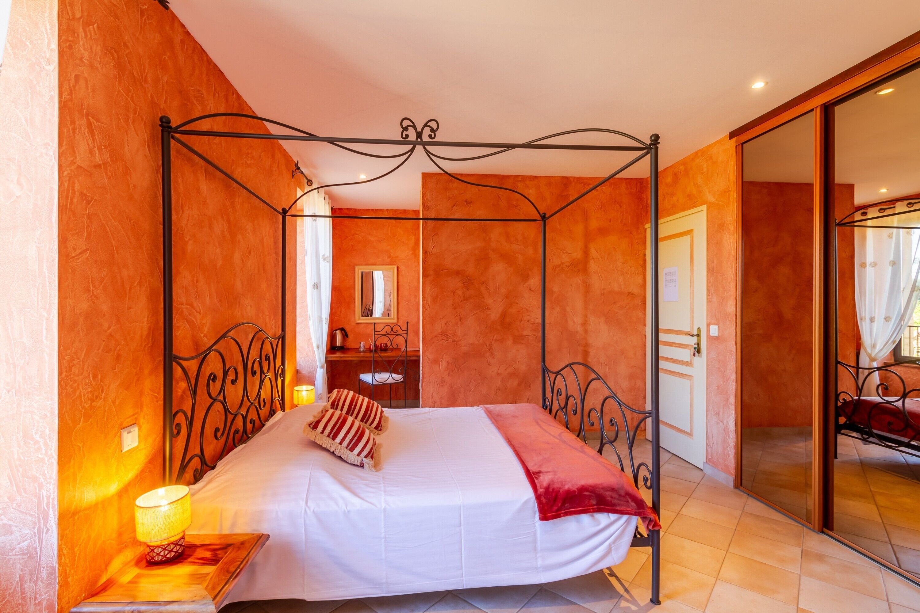 A four-poster bed with a white bedspread and red blanket, a bedside table with a lamp, and a mirror on the wall.