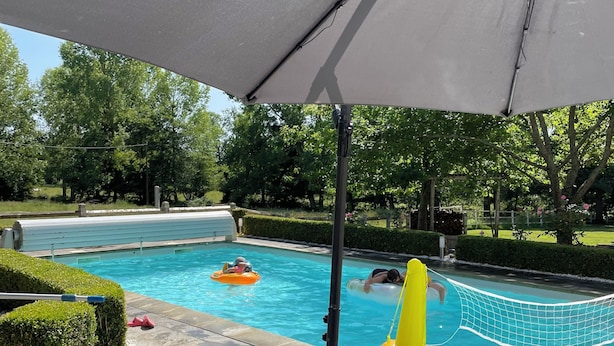 Una piscina con una red, rodeada por un solárium con baldosas y follaje.