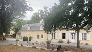 Una casa amarilla con contraventanas blancas, rodeada de árboles y plantas en macetas.