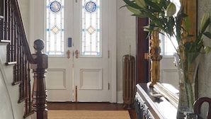 A hallway with a wooden floor, a staircase, a double door with stained glass windows, a mirror, and a vase with a plant.