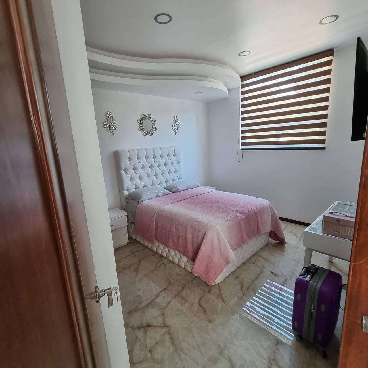 A bedroom with a tufted headboard, a bed with a pink blanket, a nightstand, a dresser, a window with blinds, and a suitcase.